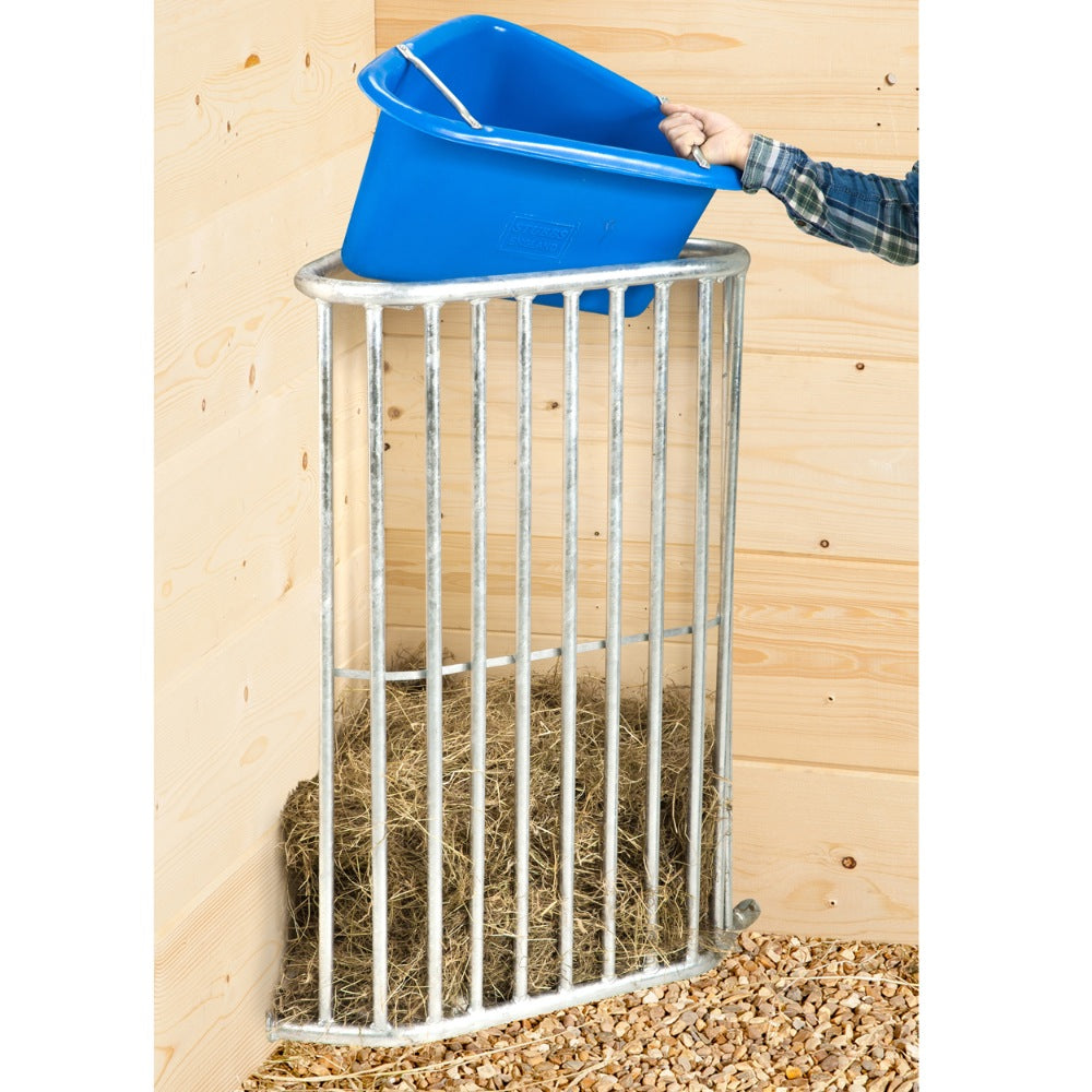 A metal feeding station with a blue tub on top, filled with hay, situated against a wooden background. Abbey England Hay Rack Available with Nice Equestrian Wukair Doha Qatar. 